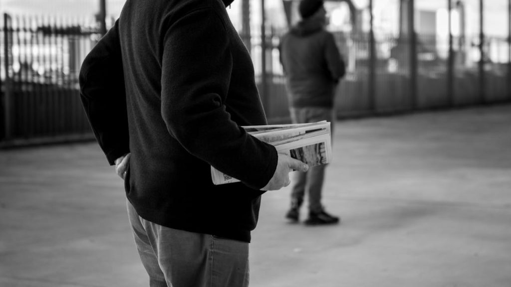 A monochrome image of a person holding newspapers in an urban environment, capturing the essence of street life.